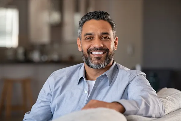 BioTE-Testosterone-Pellet-Clinic A mature South Asian man seated comfortably on a couch, smiling and looking directly at the camera satisfied with BioTE Testosterone Pellet Therapy offered by Pierre Rojas, DO in South Lyon.