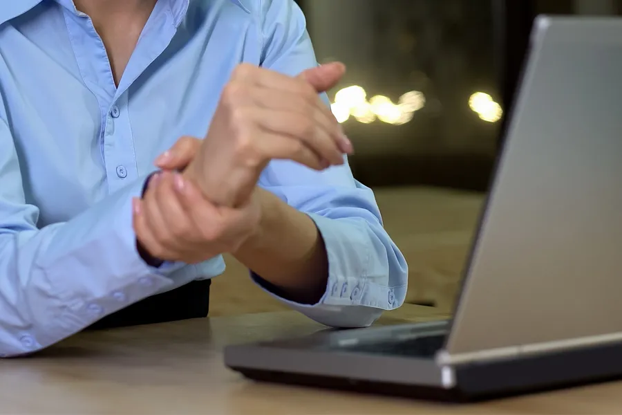 Carpal-Tunnel-Syndrome-Clinic A woman sits in front of her computer rubbing her hand in pain. Schedule treatment for carpal tunnel syndrome from Pierre Rojas, DO in South Lyon.