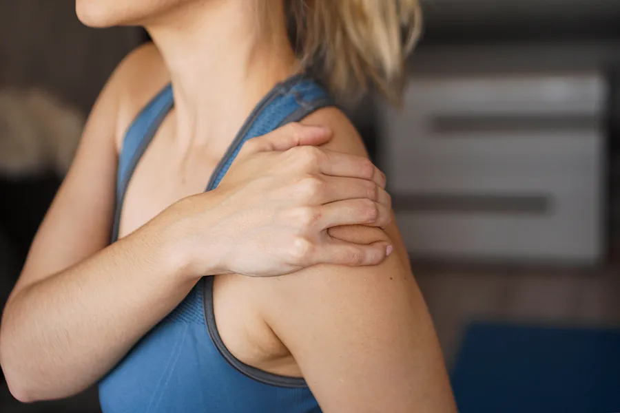 Frozen-Shoulder-Clinic A close-up view of a woman in a blue tank top, gripping her own shoulder with a look of discomfort. Receive expert Frozen Shoulder treatment by Pierre Rojas, DO located in South Lyon.