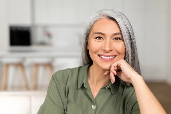 Menopause-Clinic An asian woman with long gray hair and a green shirt sitting and smiling on her couch, happy with her menopause care from [NAME} in South Lyon.