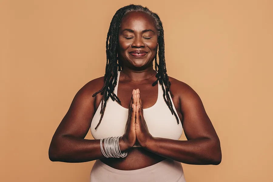 Obesity-Clinic An older African American woman confidently posing against a warm background smiling with hands in a prayer pose. Pierre Rojas, DO treats obesity using functional medicine in South Lyon.