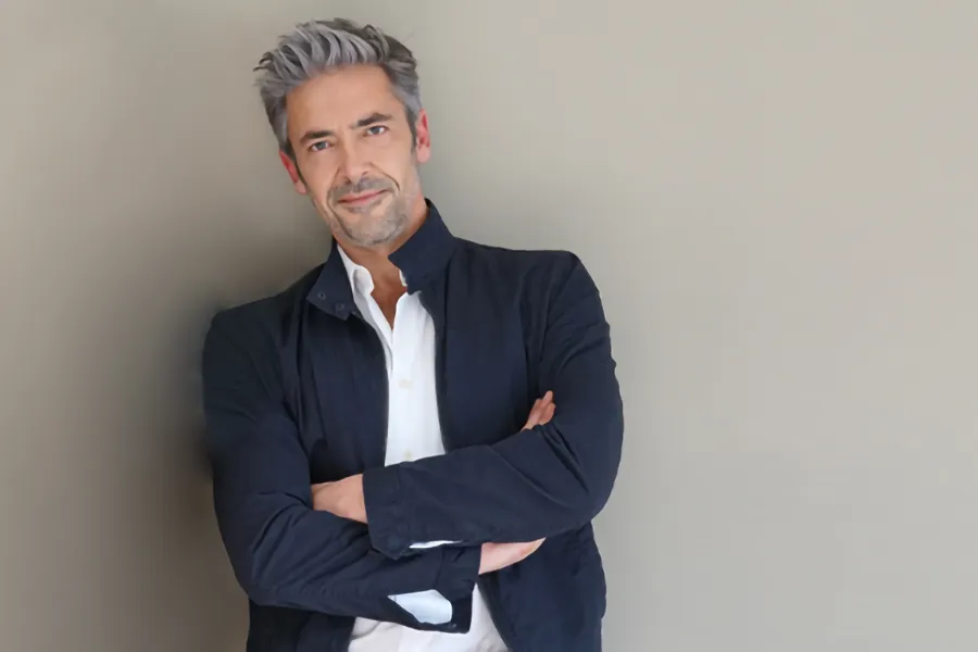 Testosterone-Low-T-Solutionsn-Clinic A middle-aged man in a white shirt and dark blue jacket leans against a gray wall smiling. Get testosterone for Low T from Pierre Rojas, DO in South Lyon.