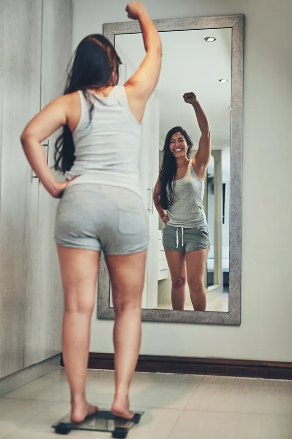 Tirzepatide-Treatment A woman in workout clothes stands on a scale looking in the mirror and pumping her hands in celebration of her successful Tirzepatide treatment from Pierre Rojas, DO in South Lyon.